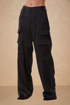 A person wearing Milan Satin Cargo Pants - Black from Noli Yoga, featuring multiple pockets, stands against a neutral background. The mid-rise cargo pants are loose-fitting and extend down to their feet. The upper torso is not visible in the image. Fall Staples, Cargo Pant, Sports Accessories, Satin Fabric, Black Pants, Jumpsuit Dress, Best Sellers, Milan, Mid Rise