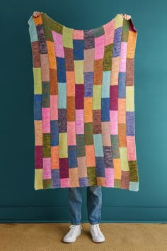 a person holding up a colorful quilt on top of a wooden floor in front of a blue wall