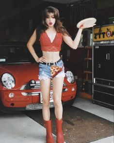 a woman standing in front of a red car holding a frisbee and wearing short shorts