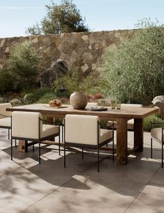 an outdoor dining table with four chairs and a bowl of fruit on the table in front of a stone wall