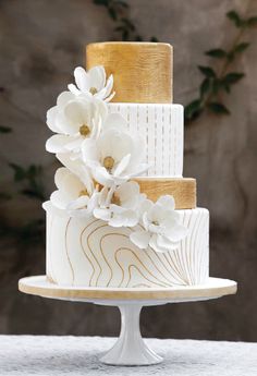 a three tiered white and gold wedding cake with flowers on the top, sitting on a table