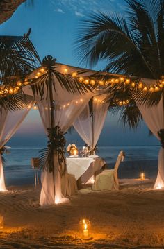 an outdoor dining set up on the beach at night with lights strung from palm trees