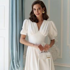 a woman in a white dress is holding a handbag and posing for the camera