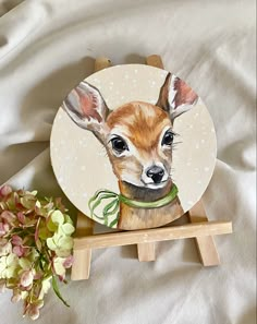 a painting of a deer on a white plate with flowers in the foreground and a wooden easel holding it