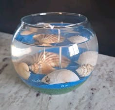 a fish bowl filled with sea shells on top of a marble counter next to a candle