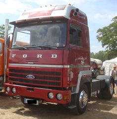 a red semi truck parked on top of a dirt field next to other trucks and people