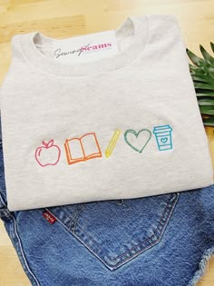 Brighten a teacher’s day with our Teacher Elements Embroidered T-Shirt! This delightful t-shirt features an array of teacher icons embroidered in the vibrant colors of the rainbow, creating a simple yet effective design that celebrates the passion and dedication of educators. Crafted from high-quality, soft, and breathable fabric, this t-shirt offers all-day comfort and durability. It’s perfect for wearing to school, casual outings, or lounging at home, making it a versatile addition to any teac Teacher Shirt Designs Ideas, Cute Teacher Sweatshirts, Teacher Hand Embroidery Designs, Fun Teacher Tshirts, Teacher Hand Embroidery, Embroidery For Teachers Gift Ideas, Embroidered Teacher Gifts, Embroidery Teacher Gifts, Teacher Inspired Outfits