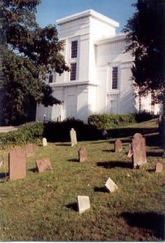 an old white church with tombstones in the front yard and trees on the other side