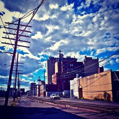 an empty street with power lines and buildings in the background under a cloudy blue sky