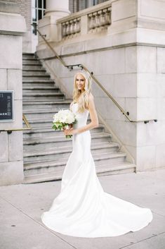 a beautiful blonde woman in a white dress holding a bouquet of flowers standing next to some stairs