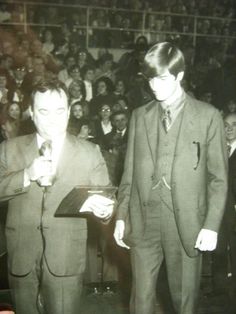 a man in a suit and tie standing next to another man holding a microphone with an audience behind him