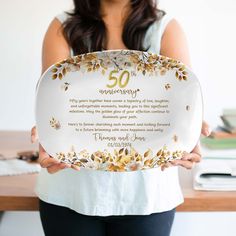 a woman holding up a 50th anniversary plate with gold flowers and leaves on the front