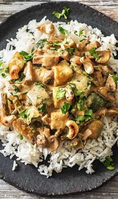 a black plate topped with white rice and meat covered in sauce on top of a wooden table