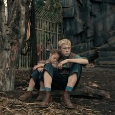 two boys sitting on the ground in front of a fence and trees with their arms around each other