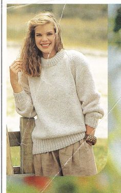 a woman wearing a white sweater and tan pants standing next to a wooden fence with her hands on her hips