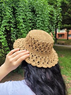 a woman wearing a brown crochet hat in front of some green trees and bushes