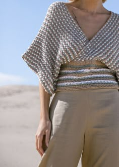 a woman standing in the sand with her hands on her hips wearing a knitted top