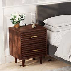 a bed with white sheets and pillows next to a night stand that has a plant on it