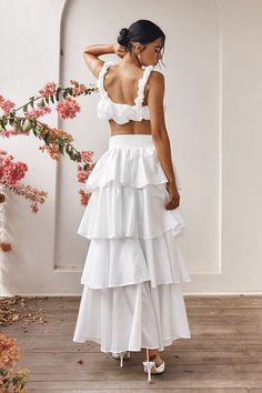 a woman wearing a white dress with ruffles on the bottom and side, standing in front of flowers