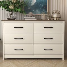 a white dresser with black handles and drawers in front of a painting on the wall