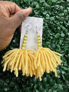 a hand holding a pair of earrings in front of some green plants with yellow beads