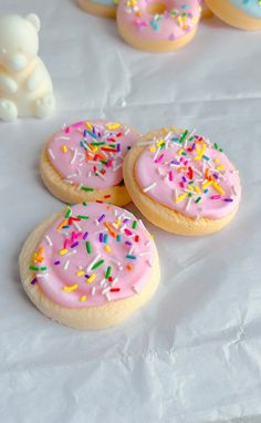 three cookies with pink frosting and sprinkles on white paper next to a teddy bear