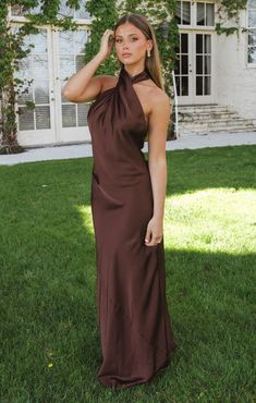 a woman standing in the grass wearing a brown dress and holding her hand to her head