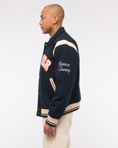 Classic bomber jacket in a soft faux wool fabric and relaxed-fit silhouette, featuring Denver Broncos varsity-inspired embroidered graphic patch details throughout, side pockets, snap-up front and tipping details at elastic cuffs and waistband. College Varsity Jacket With Padded Collar, Varsity Winter Outerwear With Padded Collar, Winter Varsity Outerwear With Padded Collar, Varsity Style Outerwear With Padded Collar For Winter, Long Sleeve Varsity Jacket With Padded Collar For Fall, Fall Varsity Sport Coat With Ribbed Cuffs, Varsity Sport Coat With Ribbed Cuffs For Fall, Winter Varsity Style Outerwear For Work, Varsity Wool Outerwear With Ribbed Cuffs