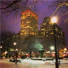 the city is lit up at night with snow on the ground and trees in front