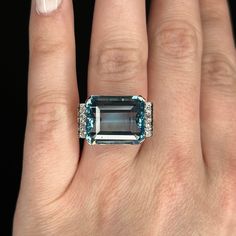 This eye-catching Art Deco cocktail ring features a 15.31 carat blue aquamarine and accent diamonds in 14 karat yellow gold and platinum. The impressive emerald cut aquamarine reflects a medium sky blue hue. Eight (8) .02 carat single cut white diamonds accent the shoulders of the band. From the profile, a retro-futuristic Art Deco basket holds the collection of gemstones finished on a platinum band. Chic, colorful, and one of a kind. Cocktail Jewellery, Aquamarine Ring Vintage, Aquamarine Cocktail Ring, Art Deco Cocktail, Art Deco Emerald Ring, Big Stone Ring, Vintage Cocktail Ring, Jewel Wedding, Cocktail Jewelry