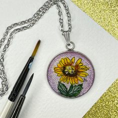 a yellow sunflower with green leaves is on a silver chain next to some paintbrushes