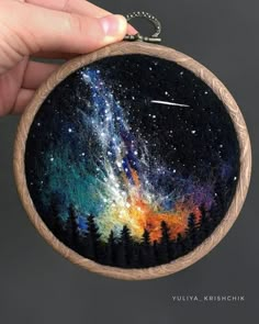 a hand holding up a small wooden hoop with an image of the night sky and stars painted on it