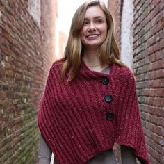 a woman standing in an alley way wearing a red knitted ponchy with buttons