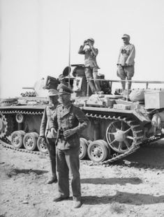 two men standing on top of a tank in the middle of dirt field next to another man