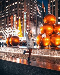 a woman standing in front of a fountain with orange balls on it and buildings behind her