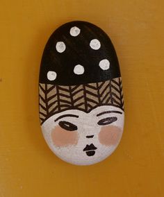 a painted rock with a woman's face on it, sitting against a yellow wall