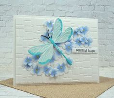 a white card with blue flowers and a dragonfly on the front, sitting against a brick wall