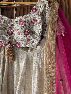 A three-piece ivory and fuchsia bridal lehenga set from the Priti Sahni collection. This beautiful georgette lucknowi thread work lehenga with a heavy border of gota, pearl, sequin, zari work border in pink accent detail is paired with a raw silk blouse with mirror, thread zardozi work all over it. The lehenga has side hanging ball tassels to the waistline. And the blouse has a sequins tassel tie-up at the back. This outfit is completed with a fuchsia net mirror butti dupatta with zari work bord Traditional Drape Lehenga With Chikankari Embroidery For Reception, Diwali Reception Lace Lehenga, Off White Embroidered Choli For Reception, Lace Choli With Intricate Embroidery For Reception, Designer Lace Lehenga For Diwali, Designer Off White Choli With Intricate Embroidery, Diwali Embroidered Lace Lehenga, Designer Bollywood Lace Choli, Off White Lehenga With Zari Work For Reception