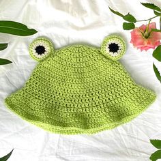 a crocheted frog hat laying on top of a white sheet next to flowers