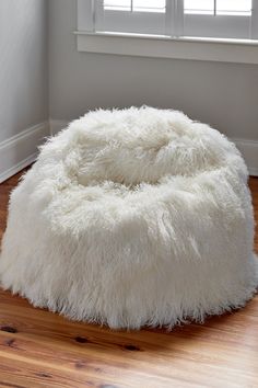 a fluffy white dog bed in the corner of a room with hardwood floors and windows