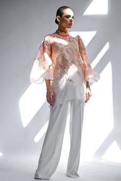 a woman standing in front of a window wearing white pants and an orange top with sequins on it