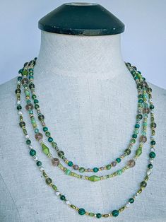 The captivating "Golden Green" necklace is a stunning combination of various beads in an array of shapes, sizes, and colors. This necklace features an eclectic mix of materials including glass, Malachite, Moss Tree Agate, Frosted Yellow Turquoise, Citrine, Purple Dyed Quartz, Diopside, Green Jade, textured and smooth golden metal, and acrylic. Each strand of this necklace is carefully crafted to showcase the unique beauty of its components adorned with a lush assortment of beads in vibrant shades of green and gold, offers a harmonious blend of textures and hues, creating a striking contrast against the skin.  Approximate length of each strand is:  - Innermost Strand 22.75 to 23.75 inches   - Middle Strand 24 to 25 inches  - Outermost Strand 27.75 to 28.75 inches  At the termination of each Green Beaded Long Crystal Necklace, Long Green Beaded Crystal Necklace, Elegant Green Beaded Long Crystal Necklace, Faceted Round Beads Necklace For Jewelry Making, Green Beaded Long Necklace With Faceted Beads, Green Long Beaded Necklaces With Faceted Beads, Bohemian Czech Glass Faceted Beads Necklace, Green Long Beaded Necklace With Faceted Beads, Green Long Necklace With Faceted Beads