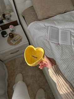 a person holding a yellow heart shaped bowl in front of a bed with an open book on it