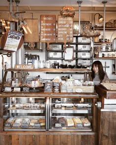 a woman standing behind a counter in a bakery with lots of food on display and signs above it