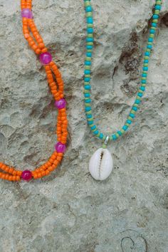 two necklaces are laying on the ground next to a seashell and an orange beaded necklace