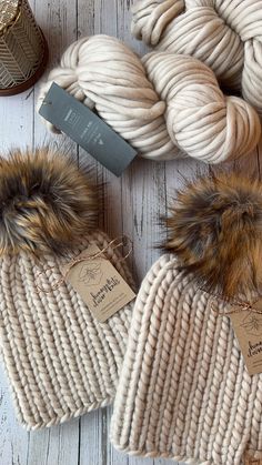 two knitted hats with fur pom - poms next to yarn and knitting needles