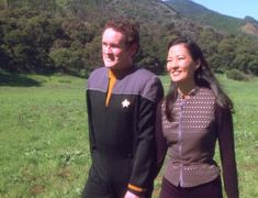 a man and woman are standing in the grass near each other, with mountains in the background