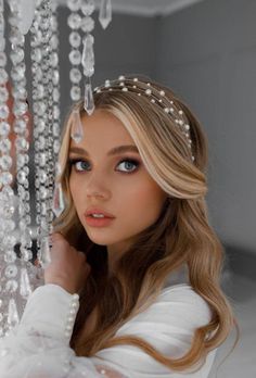 a woman with long blonde hair wearing a tiara and pearls on her head, standing in front of a chandelier