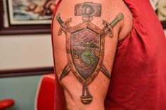 a man wearing a red shirt has a tattoo on his arm with two swords and a shield