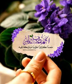 a person holding up a piece of paper with arabic writing on it and purple flowers in the background